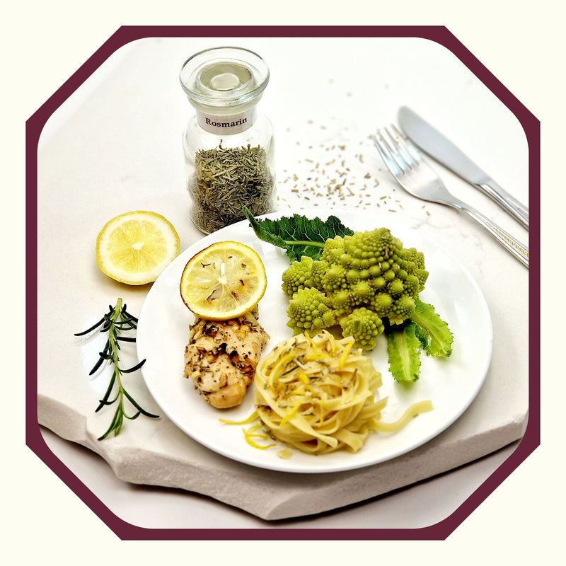 Rosmarin im Glas neben einem Teller mit einem Nudelnet, Romanesco, Rosmarin-Zitronen-Hähnchen und einer Scheibe Zitrone, einem Rosmarinzweig und Besteck