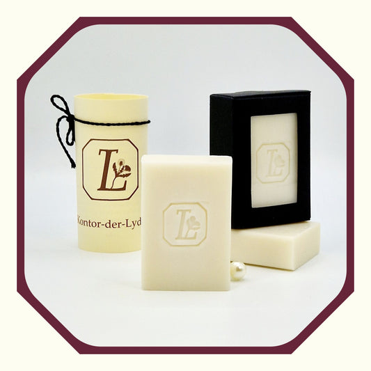 Eine Perle, eine rund gefaltete Karte mit schwarzem Schleifenband, eine Olivenseife hochkant, eine Seife schräg dahinter liegend, eine Seife Oliven-Traum hochkant auf der liegenden Seife in schwarzer Geschenkbox