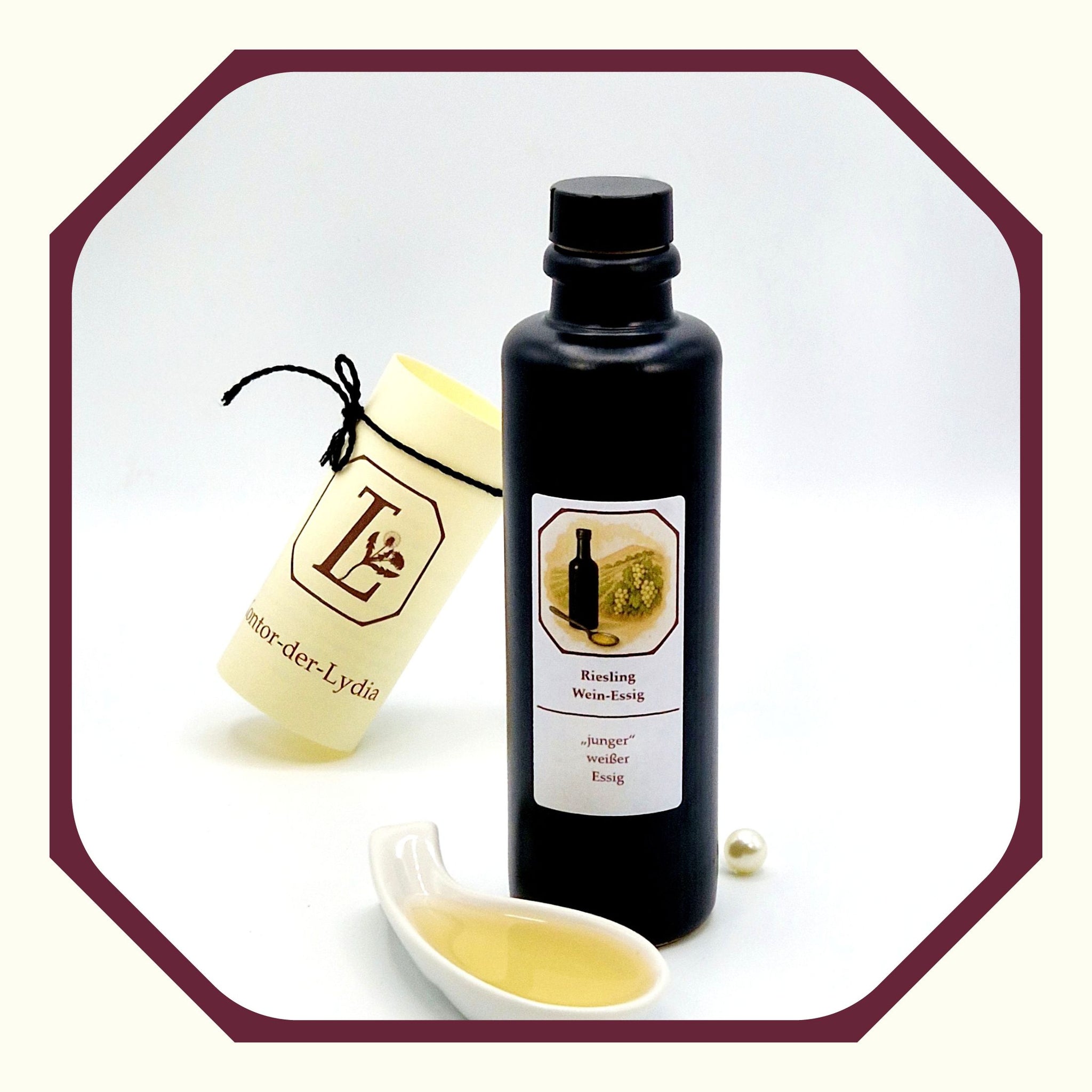 Eine Perle, eine rund gefaltete Karte mit schwarzem Schleifenband, eine schwarze Flasche mit Essig Riesling, davor eine weißer Porzellanlöffel mit Essig gefüllt