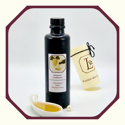 Eine Perle, eine rund gefaltete Karte mit schwarzem Schleifenband, eine schwarze Flasche mit Essig Condimento, davor eine weißer Porzellanlöffel mit Essig gefüllt