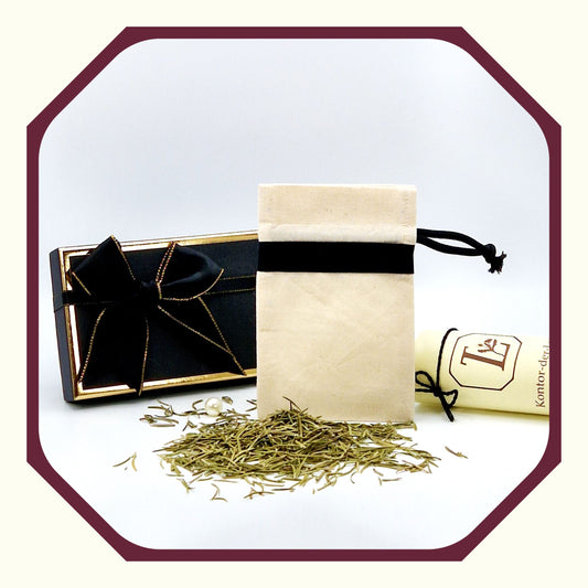 Eine Perle, eine rund gefaltete Karte mit schwarzem Schleifenband, ein Kräuter Säckchen aus Leinen vor einer kleinen Luxus  Geschenkbox, davor Rosmarin