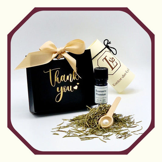 Eine Perle, eine rund gefaltete Karte mit schwarzem Schleifenband, ein Ätherisches Öl Rosmarin Premium, Rosmarin, ein Kräuterlöffel aus Holz, im Hintergrund eine kleine schwarz-goldene Geschenktasche mit dem Aufdruck Thank you und Schleife